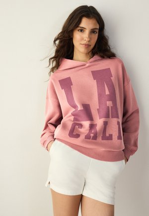 Jeune femme aux longs cheveux foncés portant un sweat à capuche rose avec le texte « LA CALI » et un short blanc, debout devant un fond clair uni.