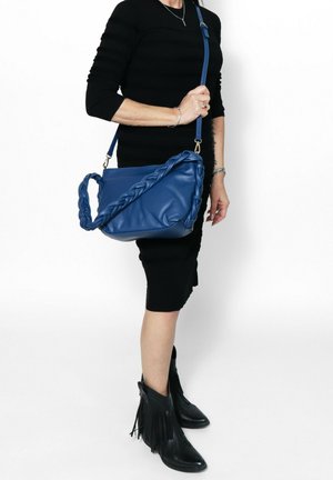 Borsa a mano blu in pelle con manico intrecciato, forma rettangolare piatta e tracolla staccabile. Texture liscia e dettagli di design minimi.