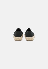 Zapatillas negras de lona sin cordones con suelas de yute trenzado y tela texturizada. Punta redondeada y diseño minimalista. Vista desde atrás.