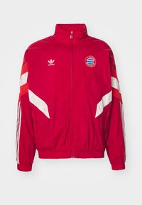 Giacca a vento rossa con accenti bianchi, zip completa, colletto alto, con logo Adidas e stemma del Bayern Monaco. Texture leggera.