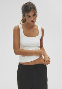 Weißes Tanktop mit quadratischem Ausschnitt, hergestellt aus glattem Stoff, kombiniert mit schwarzen Sweatpants mit Kordelzug. Verfügt über ein tailliertes Design und dezente Seitennaht.