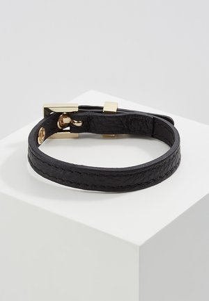 Czarna skórzana bransoletka o fakturze z pozłacaną klamrą i zapięciem, prezentowana na białym podeście na neutralnym tle.