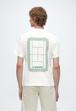 Osoba kučeravých vlasov nosí biele tričko Lacoste s grafikou zeleného tenisového kurtu a logom na chrbte, v kombinácii s béžovými nohavicami.