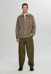 Brun fløjl zip-up jakke med krave og frontlomme, parret med olivengrønne brede bukser og sorte sko. Glat struktur.