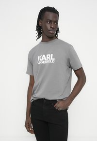 Γκρι βαμβακερή μπλούζα με έντονο λευκό λογότυπο "KARL LAGERFELD", στρογγυλή λαιμόκοψη και κοντά μανίκια. Φορεμένη με μαύρο τζιν.