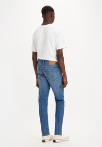 Blå denimjeans med rak skärning, med bakfickor och en läderlogotyp. Kombineras med en vit T-shirt och ljusa skor.