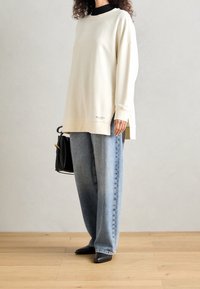 Przesadny kremowy sweatshirt z okrągłym dekoltem, bocznymi rozcięciami i detalem z logo, zestawiony z luźnymi niebieskimi dżinsami i czarnymi butami na obcasie.