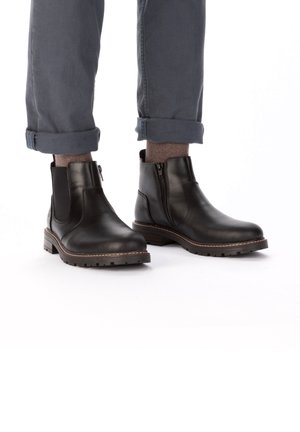Black leather chelsea boots with side zippers, rubber soles, and stitched accents, shown with rolled-up gray trousers and brown socks.