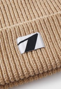 Knit beanie in tan, met ribbelstructuur. Witte label met zwart geometrisch ontwerp aan de voorkant.