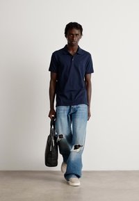 Férfi sötétkék polo pólót, világos kék, szakadt farmernadrágot és fehér sportcipőt visel, miközben sétál és egy fekete táskát tart a kezében, fehér háttér előtt.