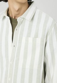 Homme portant une chemise à boutons beige et blanche à rayures verticales sur un t-shirt olive à col rond, cou et poitrine visibles.