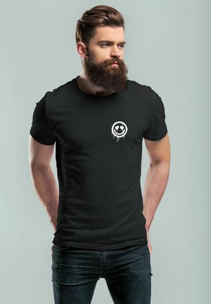 Schwarzes Baumwoll-T-Shirt mit kurzen Ärmeln, das ein weißes Tropfen-Design mit Smiley-Logo auf der Brust hat. Kombiniert mit dunklen Jeans.