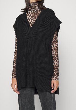 Mujer con un chaleco de punto negro sin mangas sobre un jersey de cuello alto de manga larga con estampado de leopardo y pantalones gris oscuro.