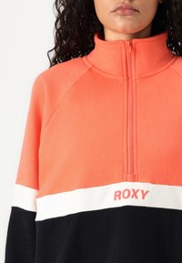 Pullover in felpa corallo con colletto alto, zip a metà, caratterizzato da righe orizzontali bianche e navy e logo "ROXY" in rosso sul petto.
