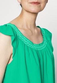 Top verde sem mangas com detalhes em babado nos ombros e um decote decorativo bordado. Tecido suave e texturizado com um corte largo.