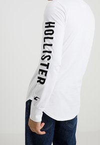 Vit långärmad skjorta med svart "HOLLISTER"-text på vänster ärm, gjord av bomullsmaterial, tillsammans med blå denimjeans.