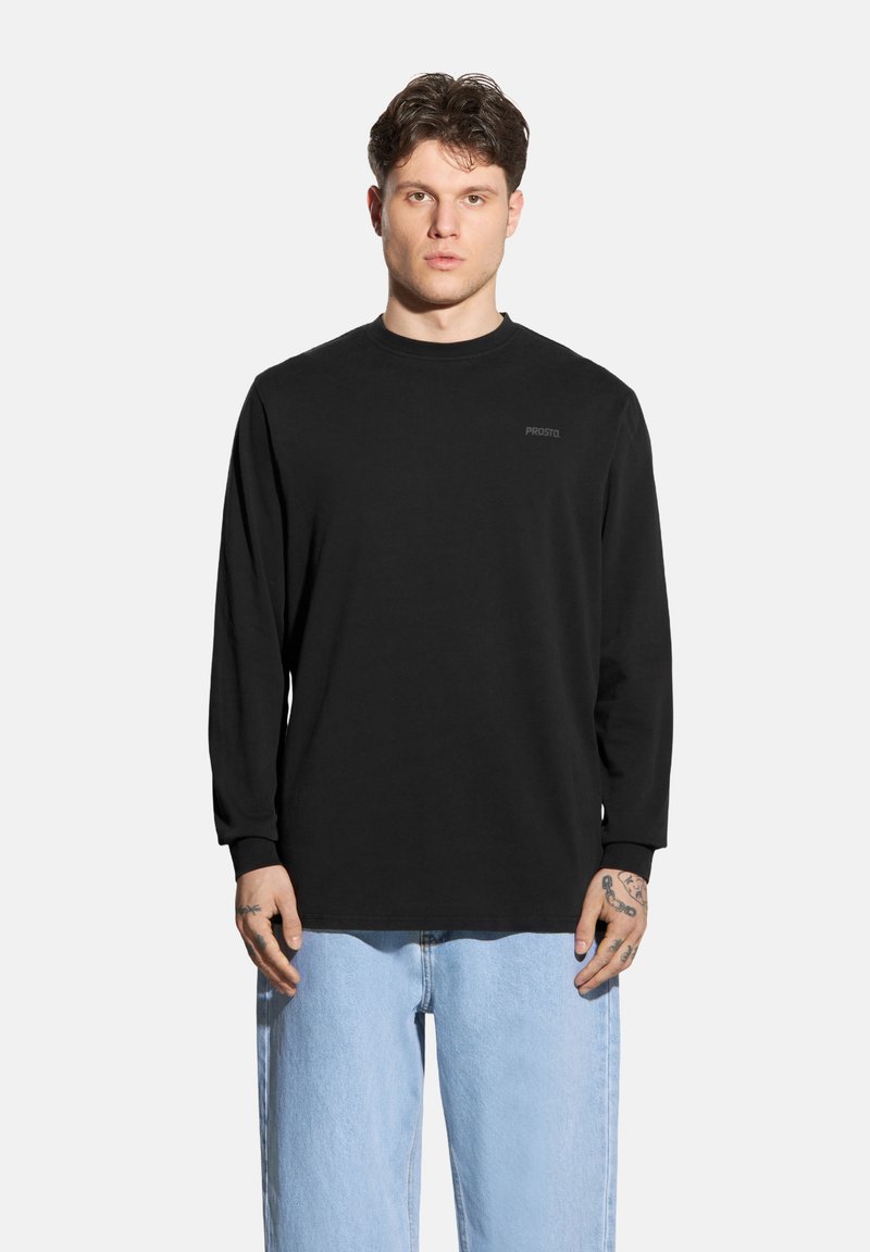 Czarna koszulka z długim rękawem o okrągłym dekolcie i subtelnym logo marki na klatce piersiowej; w połączeniu z jasnoniebieskimi, luźnymi dżinsami.