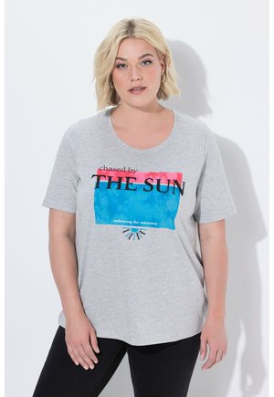 Blonde Frau trägt ein graues T-Shirt mit pinkem und blauem Schriftzug "chased by THE SUN embracing the unknown" und posiert vor weißer Wand.
