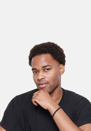 Zwarte t-shirt op een persoon met kort, krullend haar, die zijn/haar kin op hand steunt, met een dunne zwarte armband om de pols, tegen een effen witte achtergrond.