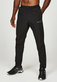 Homme athlétique portant un pantalon de survêtement noir Montebrain et des chaussures de course noires, debout les mains détendues le long du corps.