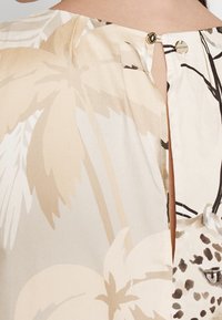 Gros plan d'un chemisier beige et crème à motifs feuilles tropicales et léopard avec un petit bouton doré au niveau de l'encolure dos.