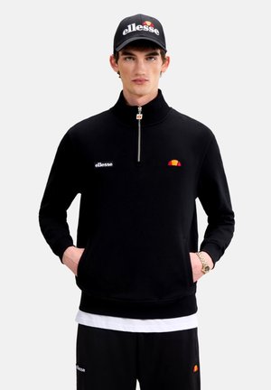 Mladý muž, ktorý má na sebe čiernu čiapku ellesse, čiernu mikinu so zipsom do polovice a čierne nohavice s logami ellesse, stojí s rukami v vreckách.