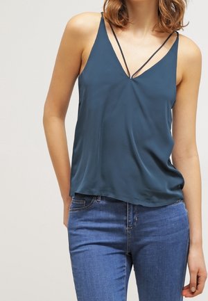 Kobieta nosząca ciemnozieloną, bezrękawnikową bluzkę na ramiączkach z podwójnymi cienkimi ramiączkami w kształcie litery V, zestawioną z wysokim stanem niebieskich dżinsów.