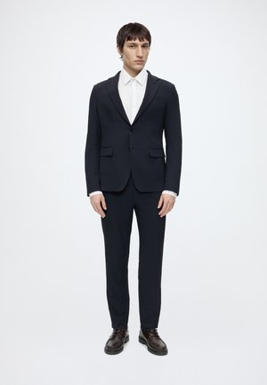 Mężczyzna stojący na białym tle, ubrany w ciemnogranatowy garnitur, białą koszulę i ciemnobrązowe eleganckie buty, zwrócony przodem, z rękami rozluźnionymi.