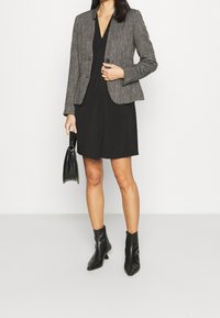 Harmaa teksturoitu bleiseri mustan mekon päällä, yhdistetty mustiin nilkkureihin ja pieneen mustaan käsilaukkuun. Yksinkertainen muotoilu, räätälöity istuvuus.