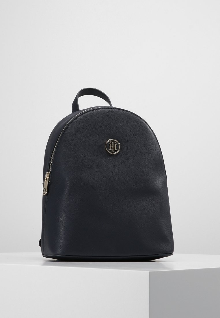 Sac à dos en cuir noir avec sommet arrondi, une seule poignée, fermeture éclair et emblème circulaire en métal avec logo sur le devant, posé sur une surface blanche.
