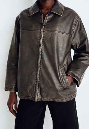 Mann trägt dunkelbraune, strukturierte Jacke mit Reißverschluss und Kragen, Hand in der Tasche, kombiniert mit schwarzen Hosen vor schlichter Hintergrund.