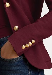 Vīna sarkans žakete ar teksturētu audumu, ar pieciem zelta dekoratīviem pogām uz aproces un roka ievietota zilā džinsa kabatā.