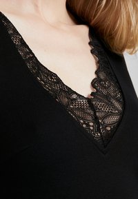 Černý oděv s hlubokým výstřihem do V, zdobený složitým černým krajkovým lemem, který ukazuje květinové vzory a vlnité okraje pro kontrast.