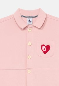 Camisa de color rosa con cuello, cuatro botones beiges y un bolsillo en el pecho que presenta un parche en forma de corazón rojo con las iniciales "PB" en blanco.
