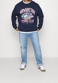 Felpa navy con grafica "MANHATTAN TENNIS CLUB", abbinata a jeans azzurri e sneakers bianche con strisce rosse.