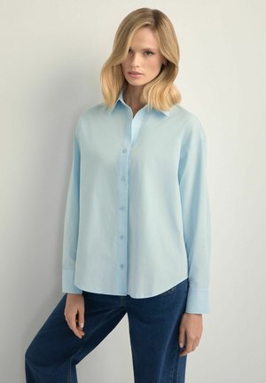 Blondynka o włosach do ramion, ubrana w jasnoniebieską koszulę na guziki i ciemnoniebieskie dżinsy, stoi na tle jednolitego jasnego tła.