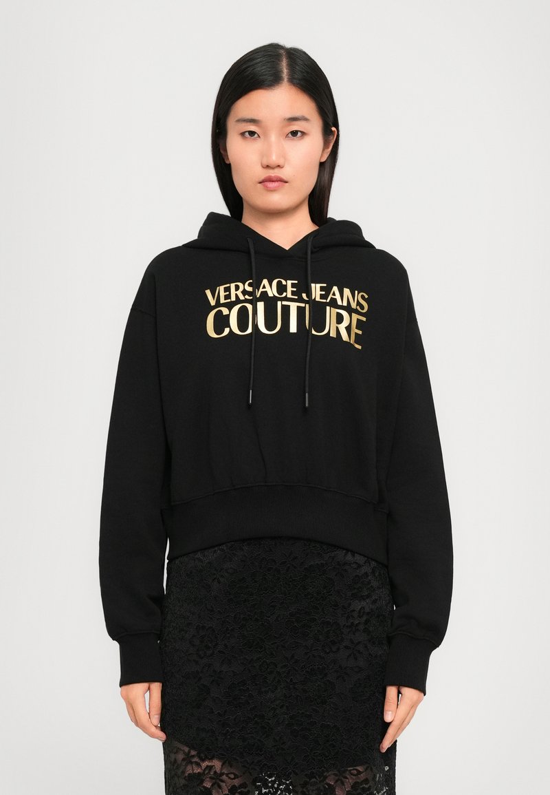Svart cropped hettegenser laget av mykt materiale med et gulltrykt logo "VERSACE JEANS COUTURE" og justerbare snører i hette.