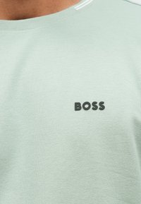 T-shirt de algodão verde claro com tecido texturizado e um logótipo "BOSS" preto na frente. Apresenta um decote redondo com riscas brancas.