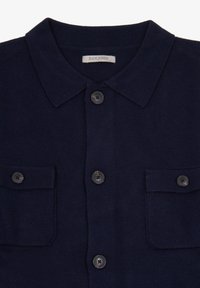Camisa de punto en azul marino con cuello abotonado, dos bolsillos frontales y botones negros. La textura suave y el diseño estructurado enfatizan la practicidad.