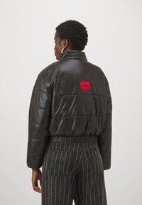 Veste cropped noire rembourrée avec une finition brillante, dotée d'un patch logo rectangulaire rouge à l'arrière et d'une taille élastiquée. Rayures verticales visibles en dessous.