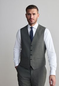 Mand med kort hår og trimmet skæg iført grå vest og bukser, hvid skjorte og mørkeblå slips, stående med den ene hånd i lommen.