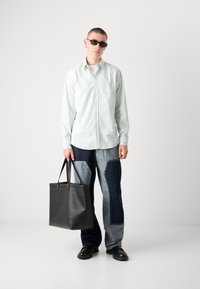 Camisa de botones verde claro, pantalones de denim azul con parches, bolso tote de cuero negro y gafas de sol. El conjunto presenta contrastes únicos de color y textura.