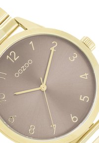 Montre-bracelet dorée avec un boîtier rond, un cadran dégradé marron, des index et des aiguilles dorées. Nom de la marque "oozoo" affiché à la position de 12 heures.