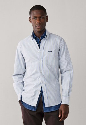 Joven con una camisa vaquera azul bajo una camisa de botones blanca y azul con rayas, posando frente a un fondo gris liso.