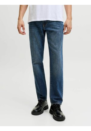 Mann iført rette blå jeans, svarte snøresko og en hvit skjorte, stående mot en ensfarget lys bakgrunn.