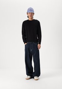 Sudadera negra, pantalones de denim oscuros y holgados, y una gorra de color azul claro. Zapatillas blancas con suelas marrones. Diseño sencillo, ajuste casual.