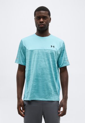 Homme debout portant un maillot de sport à manches courtes bleu clair et turquoise avec un logo, et un pantalon gris foncé, faisant face à l'avant.