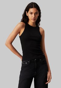 Black ribbed sleeveless top with a round neckline, paired with dark denim jeans. Features a subtle logo accent on the jeans.
