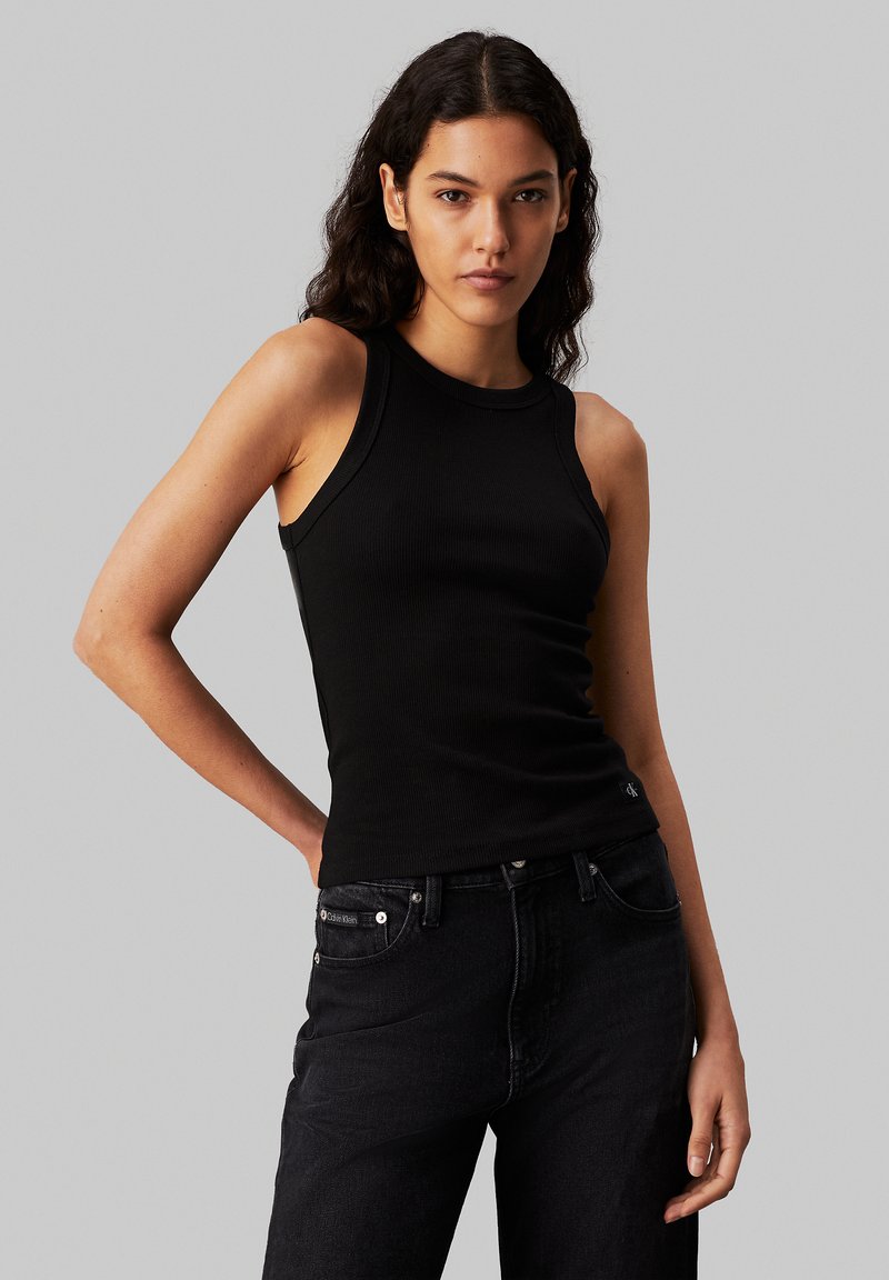 Black ribbed sleeveless top with a round neckline, paired with dark denim jeans. Features a subtle logo accent on the jeans.