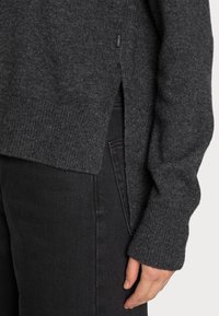 Pull en tricot gris charbon avec des poignets côtelés et des fentes sur les côtés. Le matériau semble doux avec un fini texturé. Porté sur un jean noir.